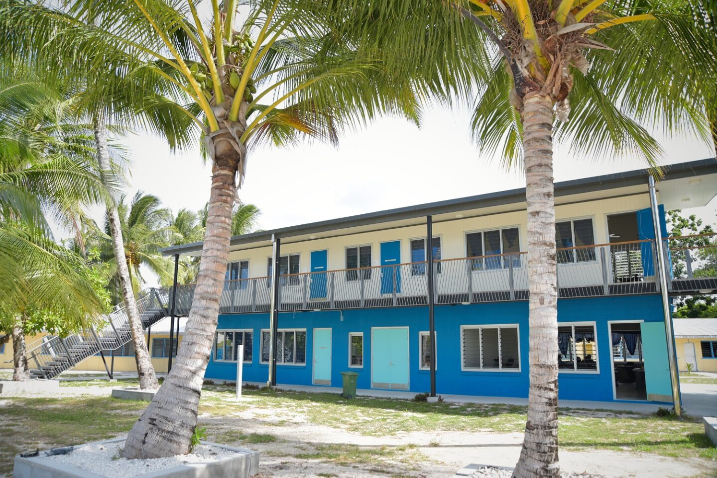 a two storey building, viewed from slightly right of the front. The bottom floor is painted blue and the top floor is painted white. There are palm trees in front of the building.